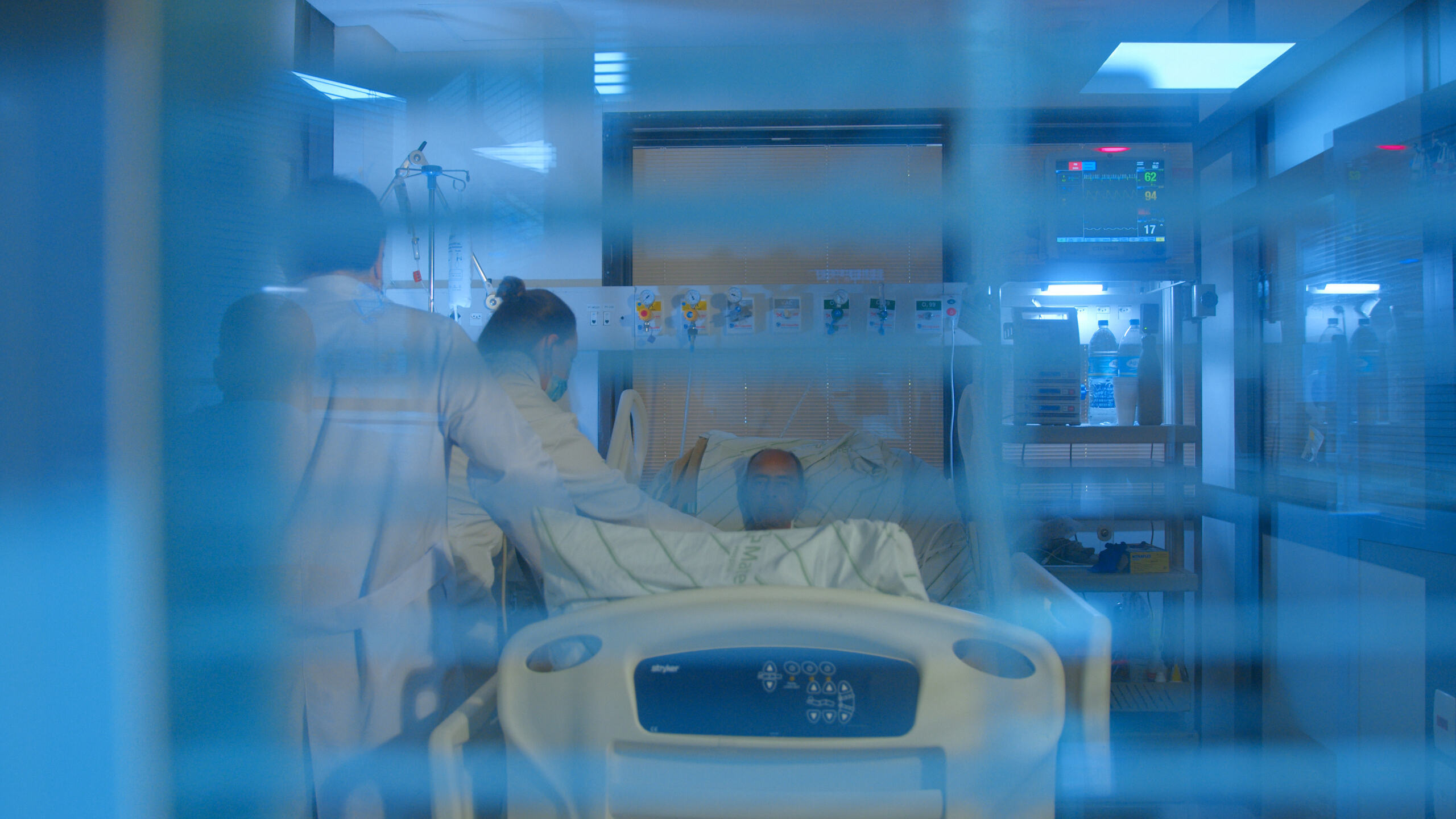 Paciente internado em leito hospitalar é atendido por profissionais de saúde em uma unidade de cuidados intensivos, com equipamentos de monitoramento ao redor.